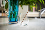 CaliWoods reusable short cocktail straw in rose gold sitting in a glass with a blue vase in the background
