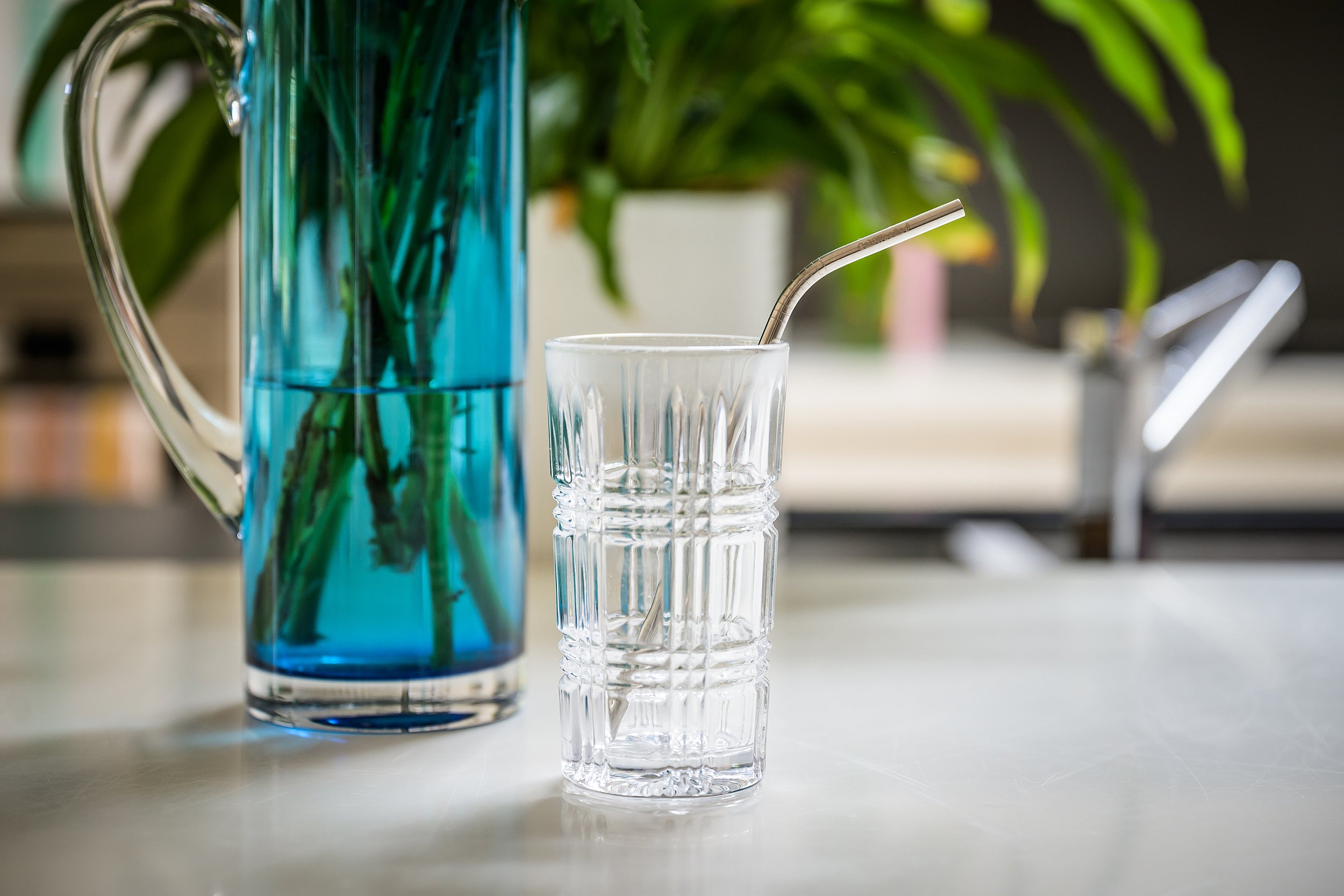 CaliWoods Reusable Drinking Straw made from stainless steel and with a bend. Straw is in a glass and there's a blue vase in the background.