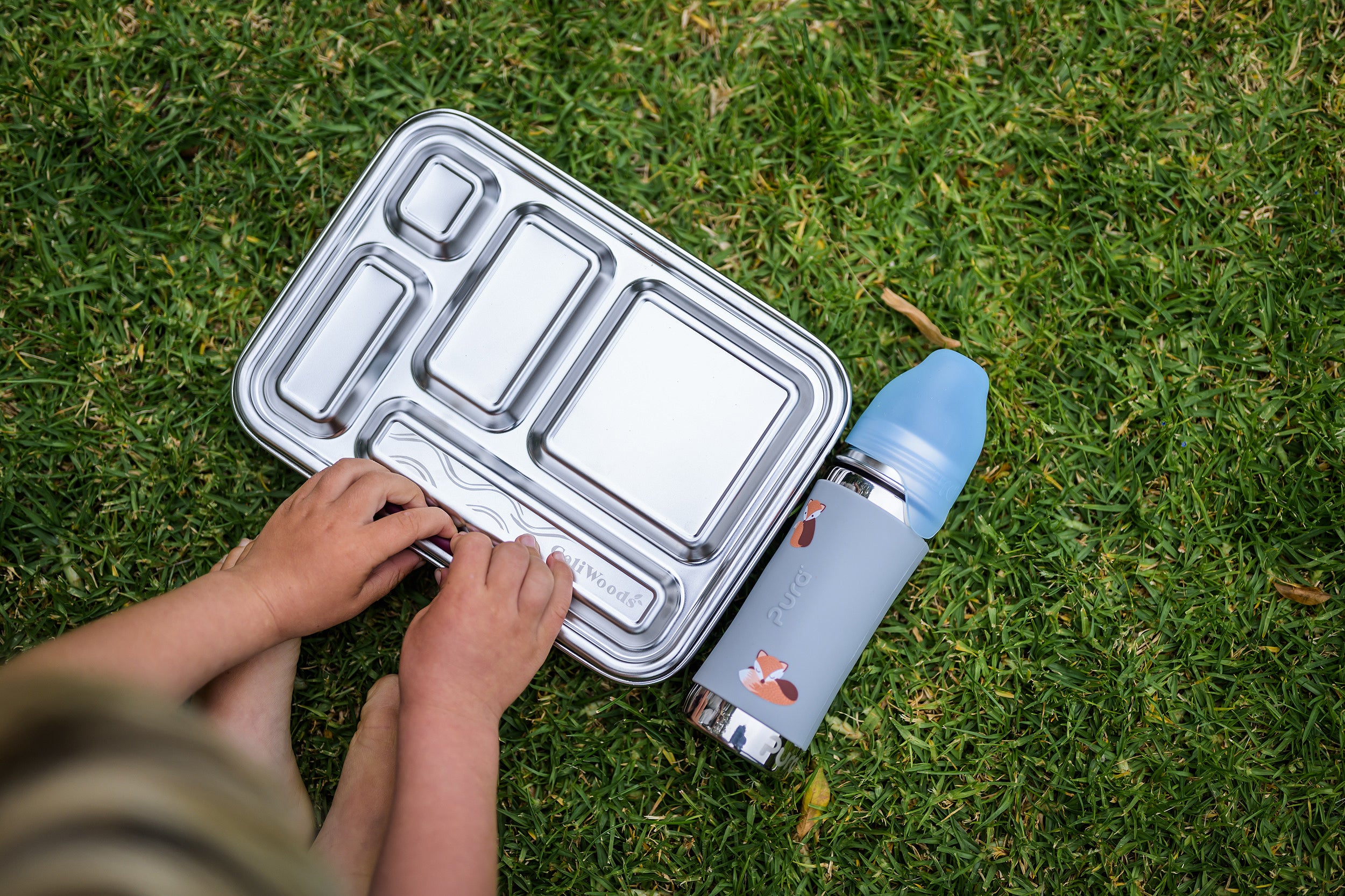 CaliWoods stainless bento toddler lunchbox on the grass with toddler hands opening it and the Pura water bottle next to it