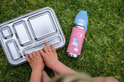 Pura Kiki toddler plastic free water bottle on the grass next to a CaliWoods stainless bento lunchbox. A little boy is using his hands to open the lunchbox 