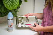 CaliWoods stainless bento lunchbox on a kitchen bench being held by a woman's hands with a stainless kid's drink bottle next to it