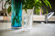 CaliWoods reusable stainless steel smoothie straw in a tall glass with blue vase in the background