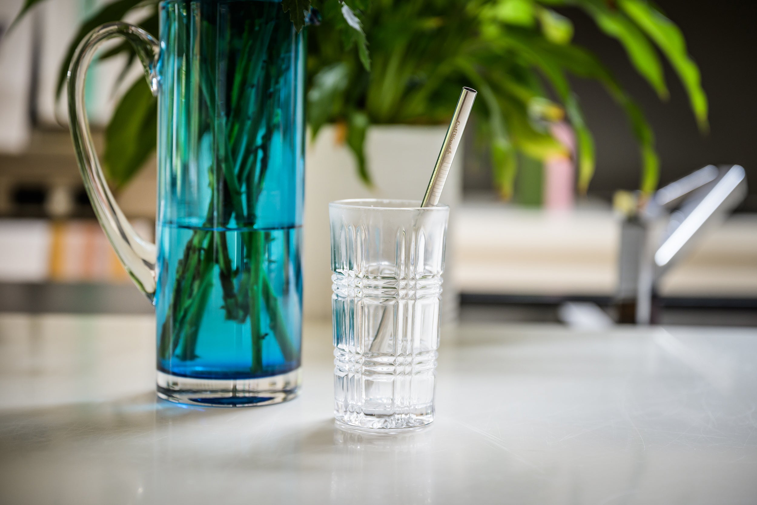 CaliWoods reusable stainless steel smoothie straw in a tall glass with blue vase in the background