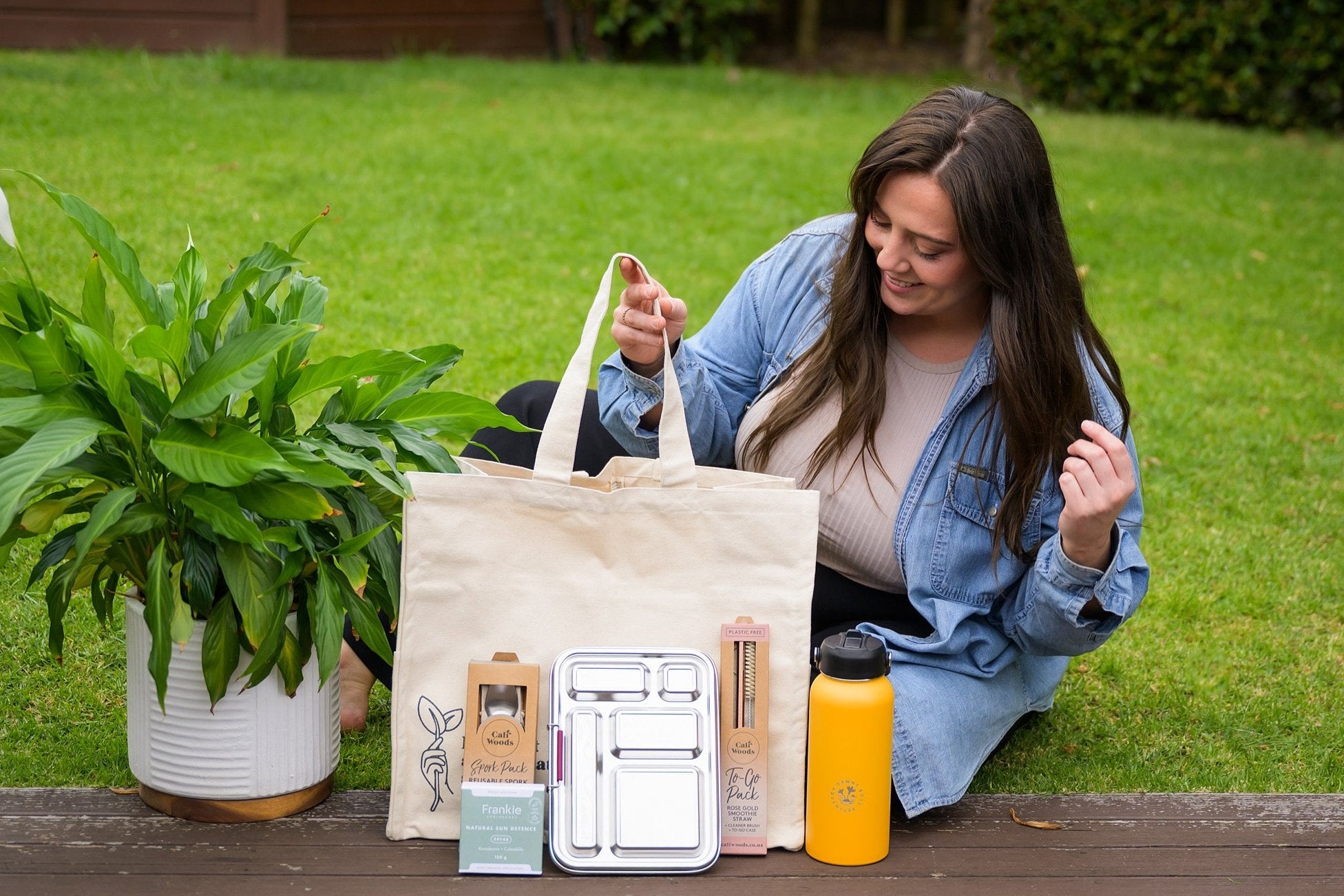 CaliWoods Summer kit with stainless lunchbox, to go straw, natural sunscreen, spork pack, dawny bottle, and tote bag held by woman sitting on the grass